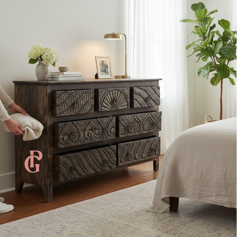 May include: A dark brown wooden dresser with six drawers, each intricately carved with floral and geometric patterns. A gold desk lamp and framed photo sit on top. A person is holding a white blanket next to the dresser.