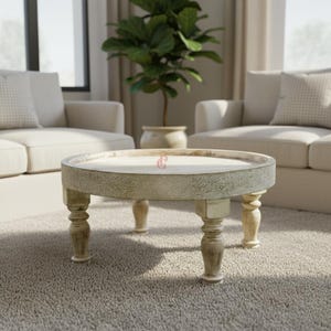 May include: A round, distressed-finish coffee table with a raised edge and four turned legs. The table is light beige and sits on a neutral-colored carpet. A potted plant and sofa are in the background.