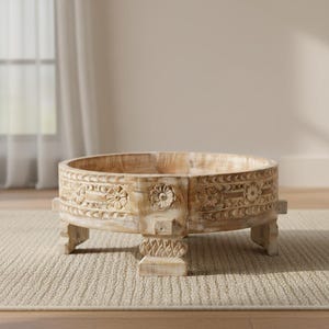 May include: A round, light-colored wooden coffee table with carved floral and geometric designs. The table has a low profile, supported by four short, square legs. The wood has a natural, distressed finish, giving it a rustic appearance.