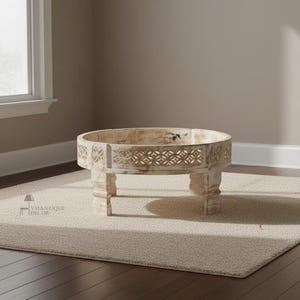 Puede incluir: Una mesa de centro redonda de madera envejecida con patrones geométricos tallados en el borde. La mesa tiene cuatro patas robustas y un acabado blanqueado, sobre una alfombra de tonos neutros. La mesa tiene unos 76 cm de diámetro.
