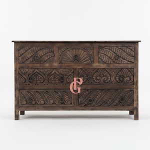 May include: A dark brown wooden cabinet with intricate carved floral and geometric patterns. The cabinet has multiple drawers and doors, with ring pulls. The design includes a sunburst motif and detailed leaf-like carvings, showcasing a rustic, handcrafted aesthetic.
