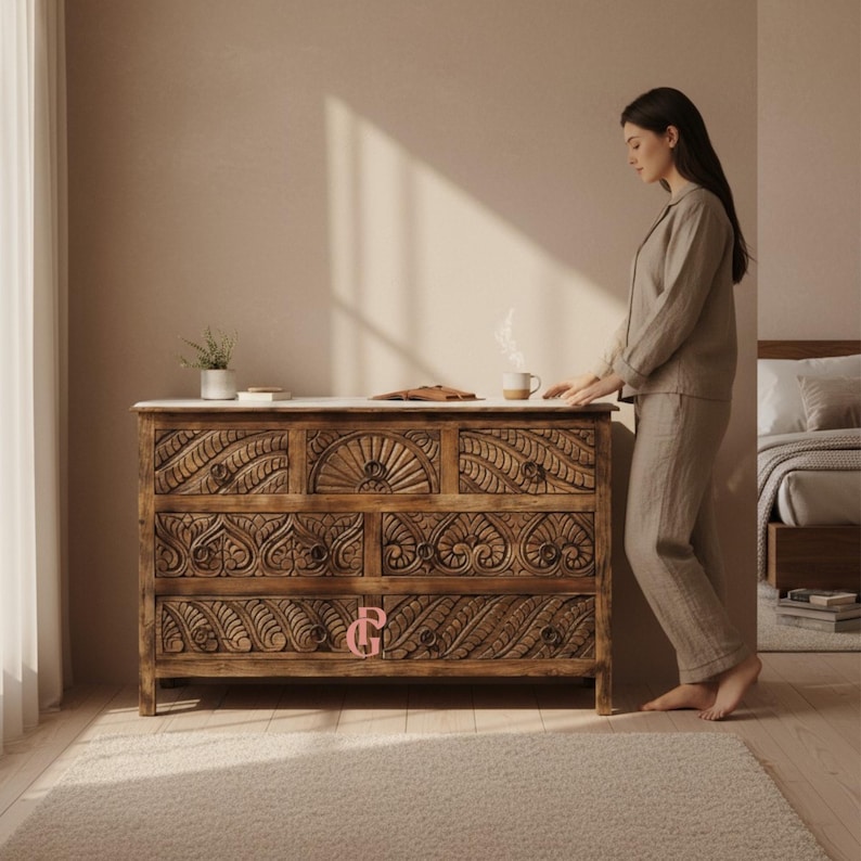 May include: A wooden dresser with intricate carved floral designs. The dresser has a marble top and multiple drawers. The wood is a warm brown color. A person is standing next to the dresser.
