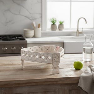May include: A distressed white wooden serving tray with a geometric design and decorative feet. The tray is placed on a wooden countertop in a kitchen setting. A green apple sits nearby.