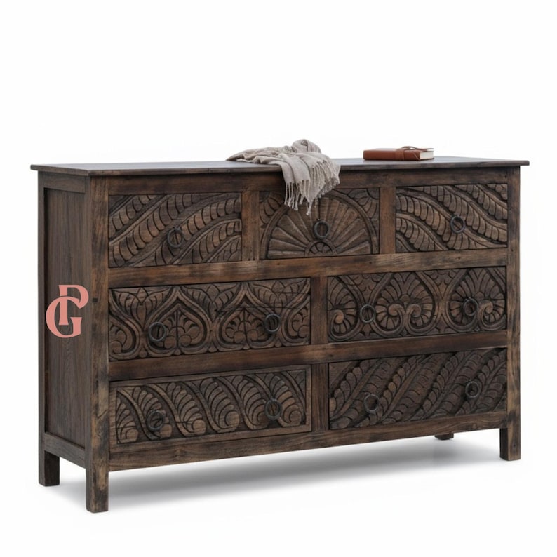 May include: A dark brown wooden dresser with seven drawers. The front of the dresser features intricate carved floral and leaf patterns. The top of the dresser has a light-colored scarf and a brown book.