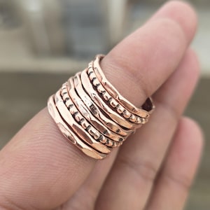 May include: A stackable copper ring with multiple bands of varying textures, including hammered and beaded designs. The ring has a warm, reddish-brown hue and is worn on a finger.