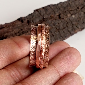 May include: A copper-colored spinner ring with three bands. The outer bands feature an ornate, etched floral design, while the central band has a textured, hammered appearance. The ring is held in a hand, with a piece of dark wood in the background.