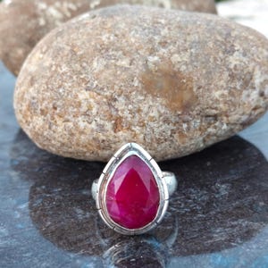 May include: A silver ring featuring a large, teardrop-shaped, faceted ruby gemstone. The ruby is a deep, vibrant red. The ring is set against a dark, reflective surface and a large, textured rock.