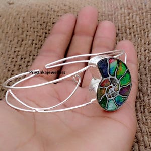 May include: A silver cuff bracelet featuring a spiral ammonite fossil centrepiece with iridescent green, red, and blue segments. The metal band is open-ended and polished. The text Priyankajewellers is visible on the side of the silver frame.