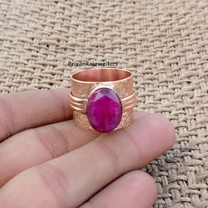 Könnte beinhalten: Ein kupferfarbener Ring mit einem großen, ovalen, rubinroten Stein. Der Ring hat ein strukturiertes Band und wird von einer Hand gehalten. Der Text "priyankajewellers" ist auf dem Ring zu sehen.