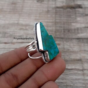 Puede incluir: Un anillo de plata con una piedra grande de color turquesa. La piedra es rectangular con una forma única y una superficie texturizada. El anillo tiene un diseño trenzado alrededor de la piedra y múltiples bandas. El texto "Priyankajewellers" es visible.