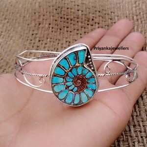 May include: A silver cuff bracelet featuring a central ammonite fossil inlay with vibrant turquoise and brown spiral patterns. The band is constructed from thin, twisted silver wire with decorative scrollwork accents. The text Priyankajewellers is visible.