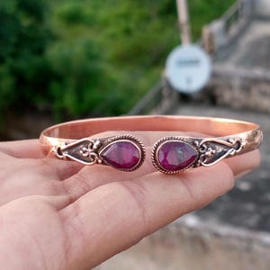 May include: A copper-colored cuff bracelet featuring two teardrop-shaped, deep red gemstones. The bracelet has ornate, dark-toned metalwork around the gemstones and along the sides. The open design allows for easy wear.