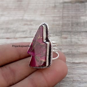 May include: A silver ring featuring a large, triangular, pink stone with gold flecks. The stone is set in a decorative silver bezel with a rope-like border. The ring has a split shank design.