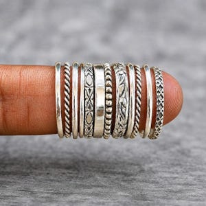 May include: A collection of eleven silver rings arranged in a row, featuring various designs including smooth bands, twisted rope textures, geometric patterns, a central polished band, and intricate floral engravings, all crafted from polished silver.