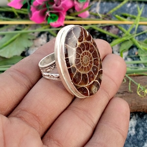 May include: A silver ring featuring an oval-shaped, polished fossil with a spiral pattern in shades of brown and tan. The ring has a wide band with decorative detailing.