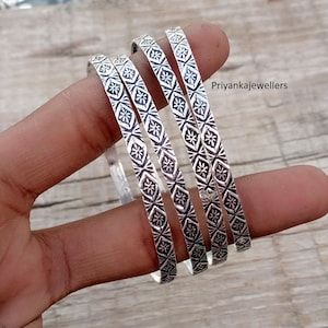 May include: Five silver bangles with a repeating geometric pattern of diamonds and floral shapes. The bangles are displayed on a wooden surface. The text "Priyankajewellers" is visible in the background.