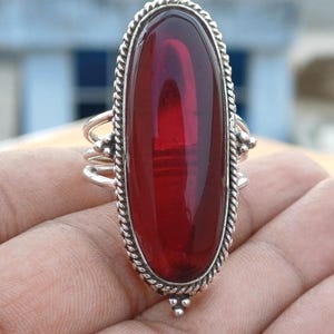 May include: A silver ring with a large, oval, deep red gemstone. The gemstone is set in a detailed silver frame with a twisted rope design around the edge. The ring has a three-band design on the sides and small decorative silver accents.