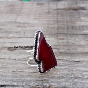 May include: A silver ring with a large, triangular, deep red stone. The stone is set in a silver band with a twisted rope design around the edges. The ring has a unique, geometric shape and is displayed on a wooden surface.