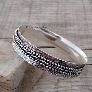 May include: A silver-colored bangle bracelet with intricate detailing. The bracelet features a textured design with multiple bands of patterns and a smooth, polished finish. The brand name "Priyankajewellers" is visible on the bracelet.