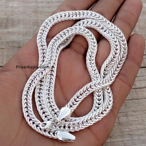 May include: A silver chain necklace with a woven pattern. The necklace is made of a shiny, metallic material and has a secure clasp. The chain is displayed on a wooden surface.
