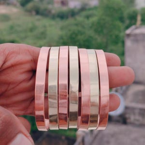 Könnte beinhalten: Eine Sammlung von Armreifen aus Kupfer und goldfarbenem Metall. Die Armreifen werden in einer Hand gehalten, wodurch ihre polierten, reflektierenden Oberflächen zur Geltung kommen. Die abwechselnden Farben erzeugen einen auffälligen Kontrast, der auf ein Schmuck- oder Accessoire-Thema hindeutet.