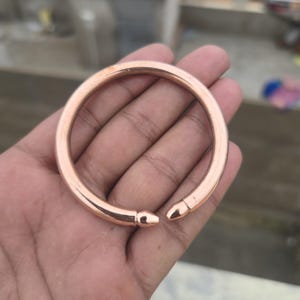 Puede incluir: Un brazalete abierto de color cobre. El brazalete tiene un diseño liso y redondeado con una ligera abertura. El metal tiene un acabado pulido que refleja la luz.