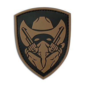 May include: A shield-shaped patch featuring a cowboy hat, two revolvers, and a masked figure. The design is primarily brown and black, with a textured border. The patch has a three-dimensional appearance.