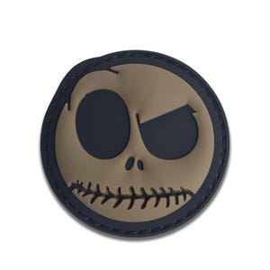 May include: A round patch featuring a stylized face with a stitched mouth and a single, angled eye. The face is tan, set against a dark blue background, and has a black border. The design is reminiscent of a popular animated character.