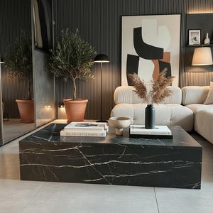May include: A modern living room featuring a large black marble coffee table with white veining. The room includes a beige sofa, a potted olive tree, and an abstract artwork. A black vase holds dried flowers.