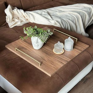 Könnte beinhalten: Ein Holzserviertablett mit goldenen Griffen hält einen weißen Gesichtstopf mit Grün, zwei Gläser und einen kleinen Holzuntersetzer. Das Tablett steht auf einem braunen Ecksofa mit einer weißen Decke.
