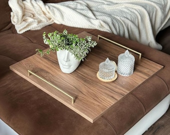 May include: A wooden serving tray with gold handles holds a white face planter with greenery, two glass jars, and a small wooden coaster. The tray sits on a brown sectional sofa with a white blanket.