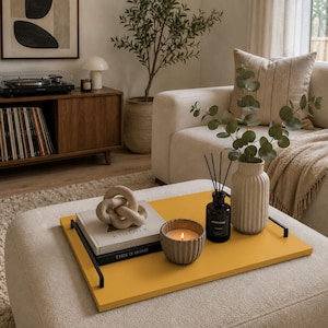 May include: A rectangular mustard yellow tray with black metal handles holds a stack of two books, a beige knot sculpture, a lit candle in a ribbed grey ceramic holder, a black glass reed diffuser bottle, and a ribbed beige vase with eucalyptus.