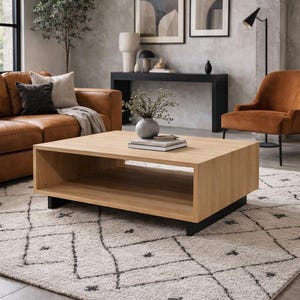 May include: A rectangular coffee table featuring a light oak wood finish with a hollow center shelf for storage. The table is supported by a black metal base and sits on a textured off-white area rug with a black geometric diamond pattern.