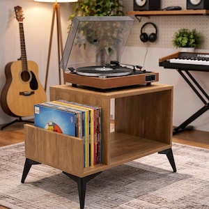 Peut inclure: Un meuble tourne-disque en bois doté d'un compartiment de rangement rectangulaire pour disques vinyles, d'une surface supérieure plane pour une platine et de quatre pieds métalliques noirs inclinés, avec une finition en bois clair.