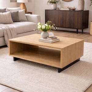 May include: A light oak rectangular coffee table with a lower shelf, supported by black feet. The table is in a living room setting with a white sofa, rug, and decorative items. A vase of flowers sits on top.