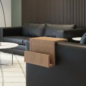 Könnte beinhalten: Ein Beistelltisch aus Holz mit einem Zeitschriftenständer, der an einer schwarzen Ledercouch befestigt ist. Der Tisch ist aus hellbraunem Holz und hat eine rechteckige Form. Im Hintergrund eine dunkelbraune Lamellenwand und ein weißer Teppich.