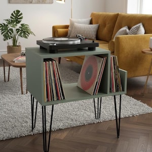 Può includere: Un mobiletto porta giradischi color verde salvia con struttura rettangolare e quattro gambe a forcina in metallo nero. La superficie superiore ospita un giradischi nero, mentre gli scomparti interni contengono numerosi dischi in vinile.
