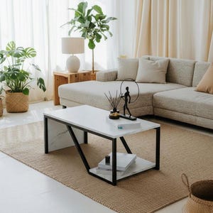 Op de afbeelding: Een moderne woonkamer met een salontafel met een wit marmeren blad en zwarte metalen poten. De tafel staat op een geweven vloerkleed, met een beige hoekbank en potplanten op de achtergrond. Een lamp en decoratieve items maken de scène compleet.