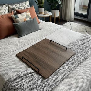 Könnte beinhalten: Ein dunkelbraunes Betttablett mit schwarzen Griffen liegt auf einem Bett mit einer weißen, gemusterten Bettdecke und einer grauen Strickdecke. Dekorative Kissen in Orange-, Grau- und Mustertönen sind am Kopfende des Bettes angeordnet.