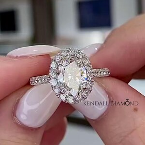 May include: A silver-toned ring featuring a central oval-cut stone surrounded by a halo of smaller round-cut stones. The band is encrusted with tiny pavé-set stones. The text KENDALL DIAMOND is visible on the ring setting near the center stone.