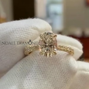 May include: A gold engagement ring with a large oval-cut diamond center stone. The band is also adorned with smaller diamonds. The ring is held by a gloved hand, with the text "KENDALL DIAMOND" visible.