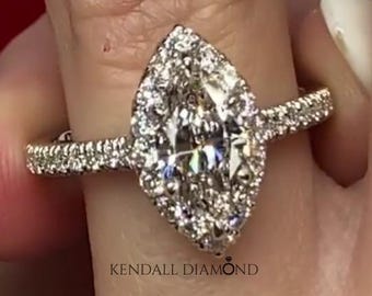 Könnte beinhalten: Ein Ring mit einem Diamanten im Marquise-Schliff, umgeben von einem Halo aus kleineren runden Diamanten, auf einer dünnen Ringschiene mit Pavé-Diamanten. Die Ringschiene ist silberfarben und der Text KENDALL DIAMOND ist zu sehen.