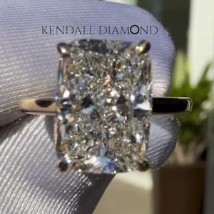 Könnte beinhalten: Ein Ring mit einem Diamanten im Radiant-Schliff, der einen großen, rechteckigen Mittelstein mit zahlreichen lichtreflektierenden Facetten aufweist, gefasst in eine dünne, polierte Metallschiene. Der Text KENDALL DIAMOND ist über dem Ring zu sehen.