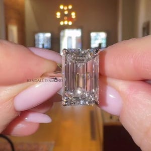 May include: A clear, rectangular-cut diamond ring is held up. The diamond has many facets and sparkles. The ring is set in a silver band. The text "KENDALL DIAMOND" is visible on the ring. The background is blurred.