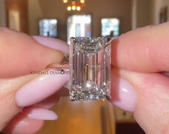 Op de afbeelding: Een heldere, rechthoekig geslepen diamanten ring wordt vastgehouden. De diamant heeft veel facetten en schittert. De ring is gezet in een zilveren band. De tekst "KENDALL DIAMOND" is zichtbaar. De achtergrond is wazig.