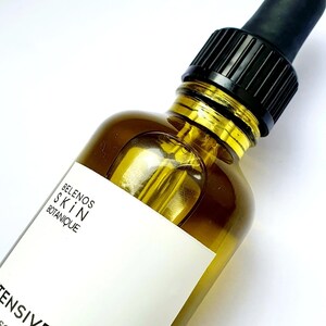 May include: A clear glass bottle of skincare product with a black dropper. The bottle contains a golden-colored liquid. The label reads "BELENOS SKIN BOTANIQUE" and "INTENSIVE ELIXIR". The product is designed to encourage healthy and hydrated skin.