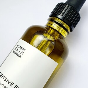 May include: A close-up of a skincare product bottle. The bottle is olive-green glass with a black dropper and cap. A white label displays the brand name and product description. The bottle is filled with a golden liquid.