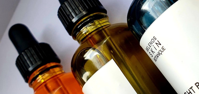 May include: Close-up of three amber glass bottles with black droppers. The bottles contain a clear, golden liquid. One bottle has a white label with the text "Belenos Skin Botanique".