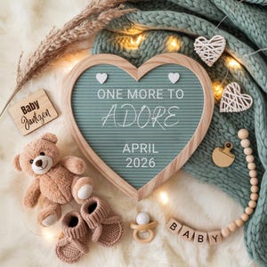 Peut inclure: Une annonce de grossesse avec un tableau en forme de cœur affichant "ONE MORE TO ADORE APRIL 2026". L'image comprend un ours en peluche, des chaussons de bébé, une tétine et des blocs en bois épelant "BABY". Des cœurs décoratifs et une couverture en tricot complètent la scène.