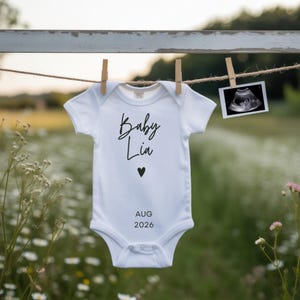 Könnte beinhalten: Ein weißer Baby-Body mit dem Schriftzug "Baby Lia" in Schreibschrift, einem schwarzen Herz und dem Datum "AUG 2026" darunter. Der Body hängt an einer Wäscheleine mit einem Ultraschallbild daneben.